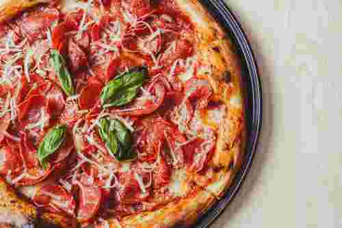 Das Bild zeigt eine frisch gebackene Pizza mit Salami, geriebenem Käse und Basilikumblättern auf einem dunklen Pizzateller.
