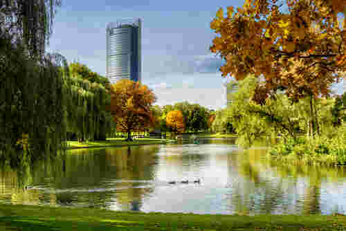 Städtischer Park mit Teich, buntem Herbstlaub und schwimmenden Enten, im Hintergrund ein moderner Hochhausturm vor blauem Himmel.