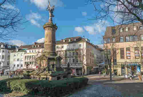Historischer Platz mit steinernem Denkmal und Siegesfigur, umgeben von Hotels, Geschäften und Passanten bei blauem Himmel und Sonnenlicht. Historischer Platz mit steinernem Denkmal und Siegesfigur, umgeben von Hotels, Geschäften und Passanten bei blauem Himmel und Sonnenlicht.