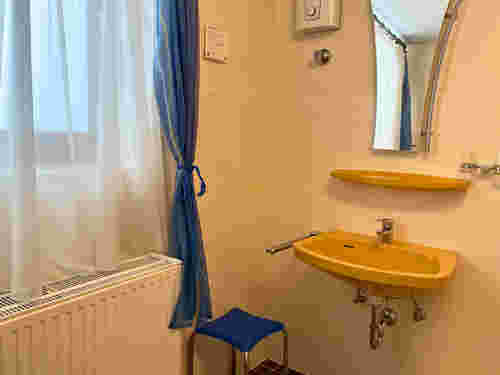 Gelbes Waschbecken mit Ablage und Spiegel, daneben ein Fenster mit blau-weißer Gardine und ein blauer Hocker im Bad. Gelbes Waschbecken mit Ablage und Spiegel, daneben ein Fenster mit blau-weißer Gardine und ein blauer Hocker im Bad.