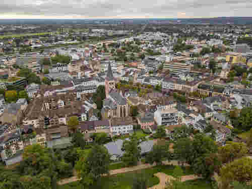 Luftaufnahme einer dicht bebauten Innenstadt mit markanter Kirche im Zentrum, umgeben von Wohn- und Geschäftsgebäuden sowie viel Grün im Vordergrund. Luftaufnahme einer dicht bebauten Innenstadt mit markanter Kirche im Zentrum, umgeben von Wohn- und Geschäftsgebäuden sowie viel Grün im Vordergrund.