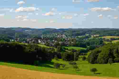 Blick über eine grüne Hügellandschaft auf ein Dorf mit Kirche im Zentrum, umgeben von Wäldern, Feldern und blauem Himmel mit Wolken.