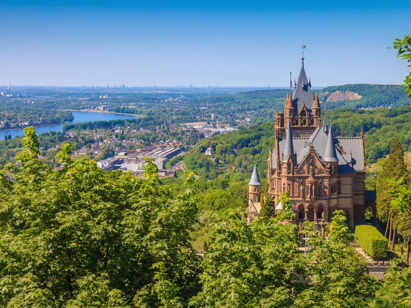 Blick auf das Schloss Drachenburg mit seinen Türmchen, umgeben von grünem Wald, im Hintergrund der Rhein und die weite Rheinebene bei klarem Himmel.