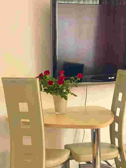 Kleiner Tisch mit zwei Stühlen beim Wandfernseher mit Rosen auf dem Tisch Kleiner Tisch mit zwei Stühlen beim Wandfernseher mit Rosen auf dem Tisch