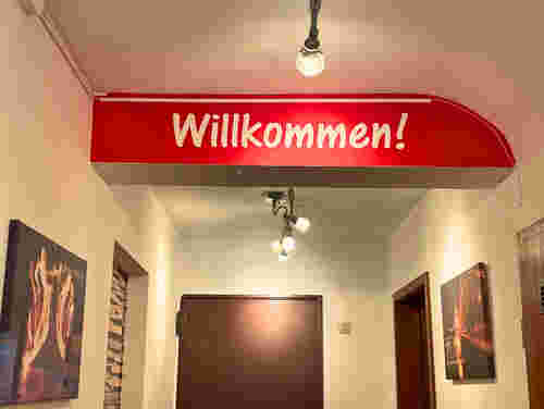 Flur mit weißer Wand, roten Akzenten und großem „Willkommen!“-Schriftzug, beleuchtet durch mehrere Deckenstrahler, mit Wandbildern.