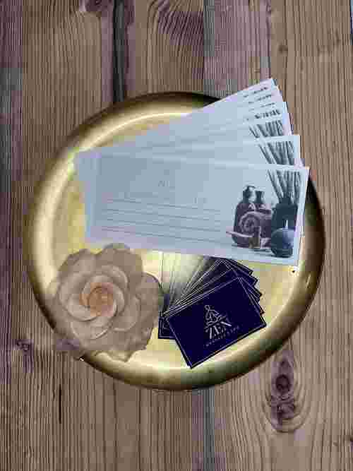 Das Bild zeigt dekorative Geschenkgutscheine und Visitenkarten vom „ZEN Massage & Spa“ auf einem goldfarbenen Tablett, arrangiert mit einer zarten Stoffblume. Das Bild zeigt dekorative Geschenkgutscheine und Visitenkarten vom „ZEN Massage & Spa“ auf einem goldfarbenen Tablett, arrangiert mit einer zarten Stoffblume.