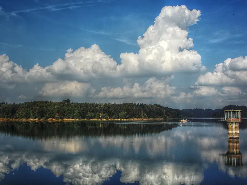 Stausee mit bewaldetem Ufer, markantem Pegelhaus und spiegelglatter Wasseroberfläche, darüber eindrucksvolle, hohe Quellwolken vor blauem Himmel.