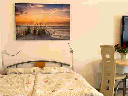 Schlafzimmer mit Doppelbett und gemusterter Bettdecke, darüber ein Bild mit Sonnenuntergang am Strand; daneben Tisch mit Vase und Rosen. Schlafzimmer mit Doppelbett und gemusterter Bettdecke, darüber ein Bild mit Sonnenuntergang am Strand; daneben Tisch mit Vase und Rosen.